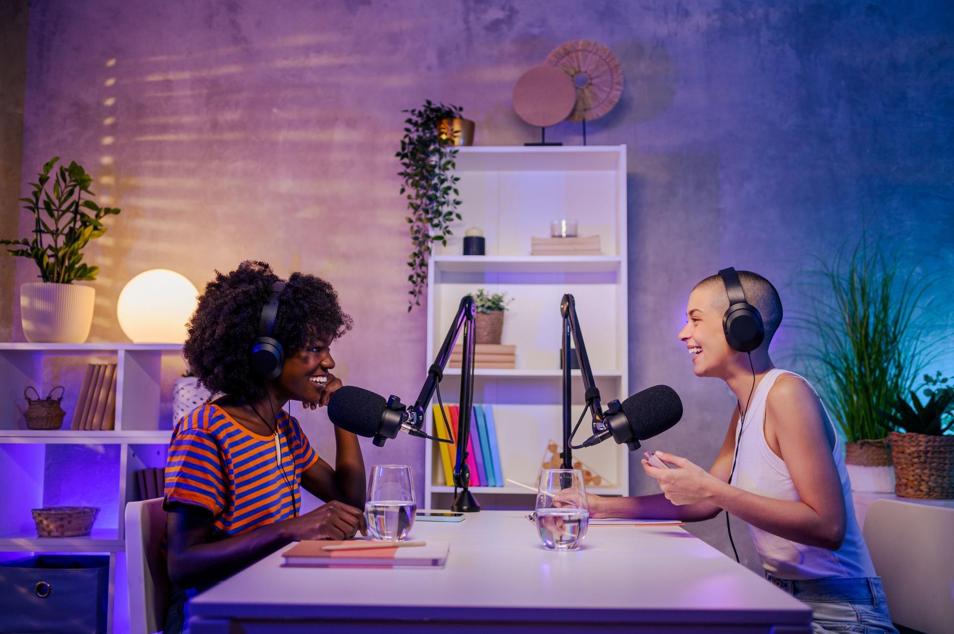 Two interracial podcasters are sitting in a home broadcasting studio and discussing topics while talking at the microphone.